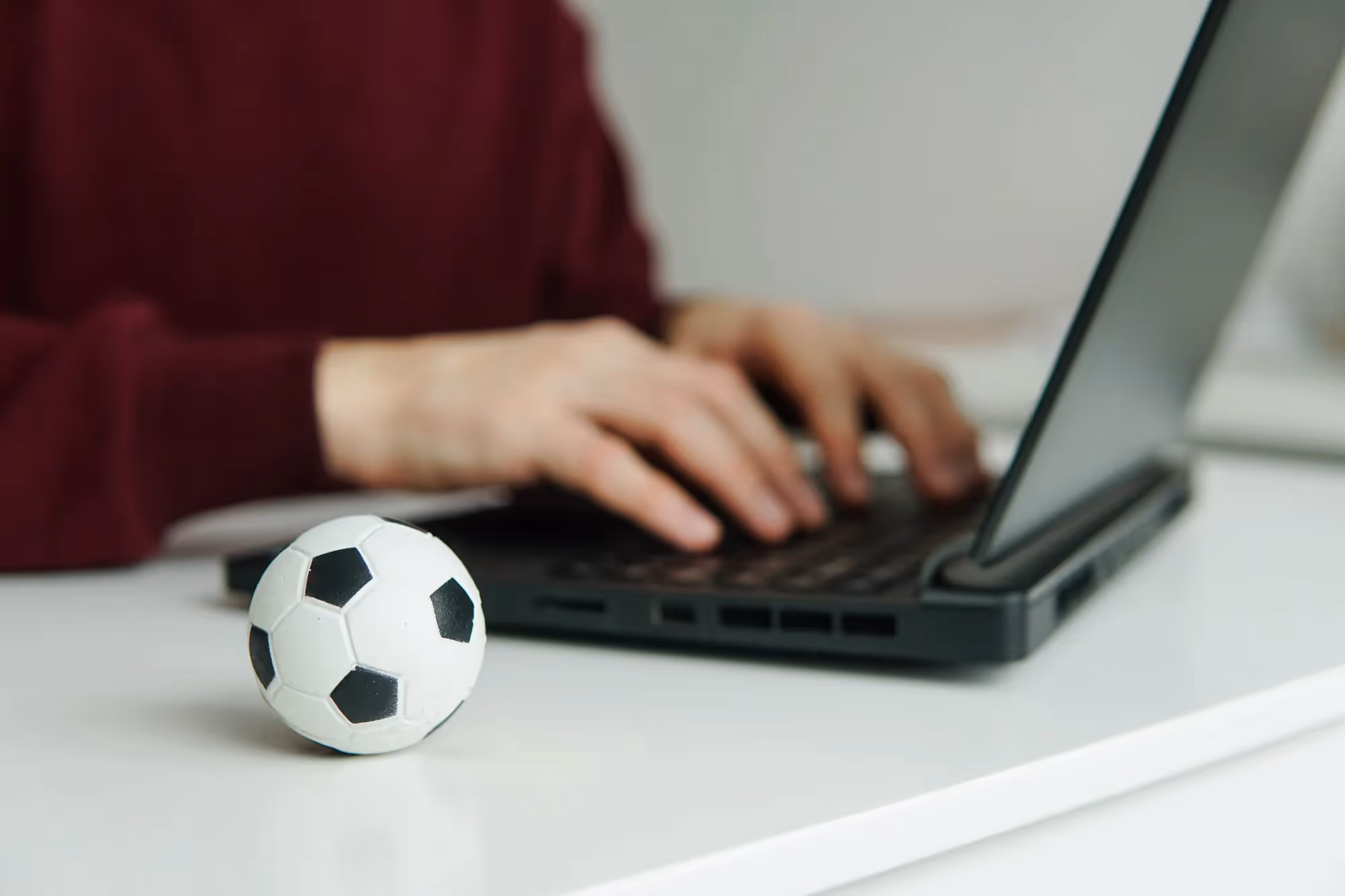 A man is typing on a laptop with a soccer ball on the desk.