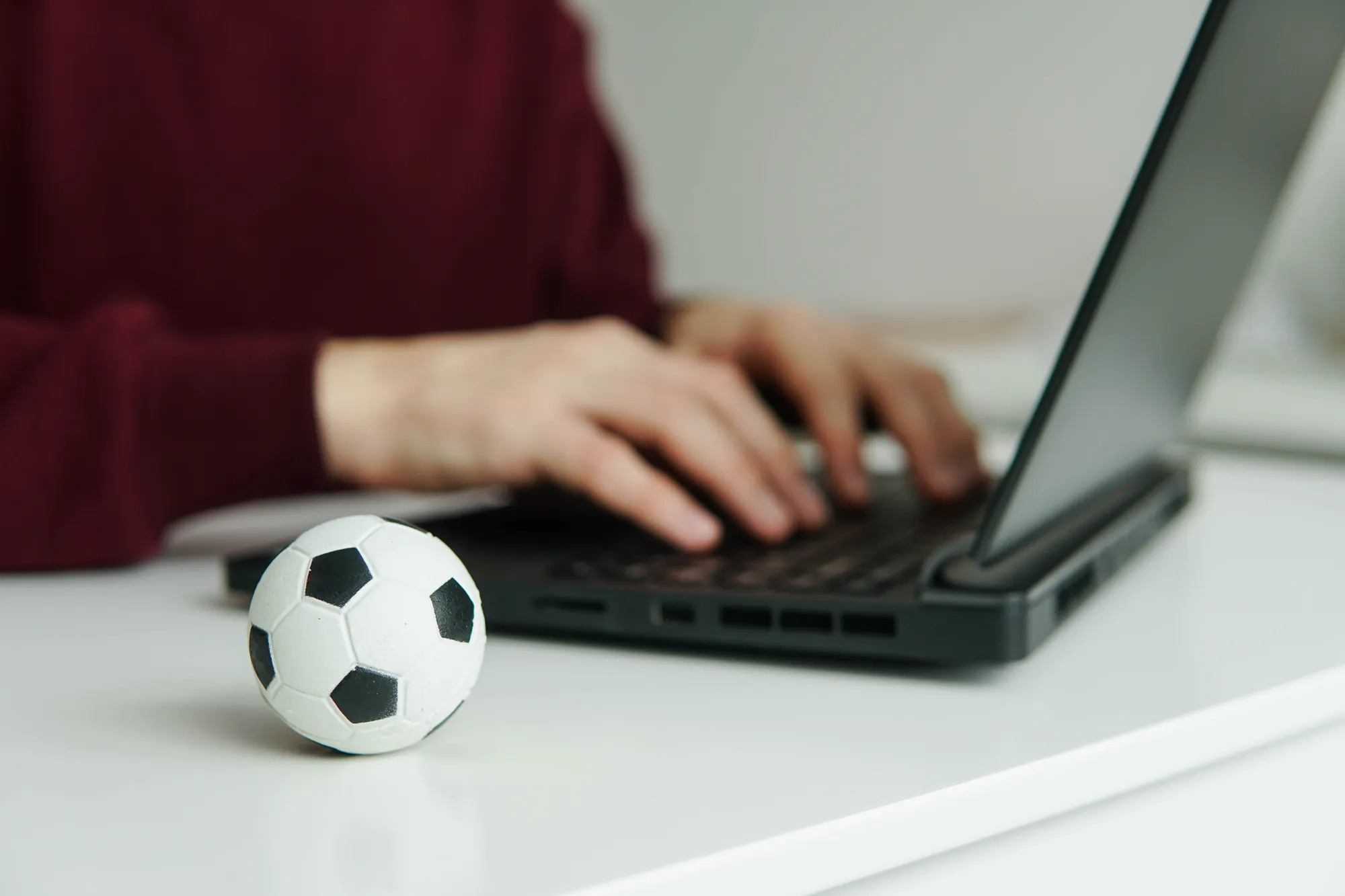 A man is typing on a laptop with a soccer ball on the desk.