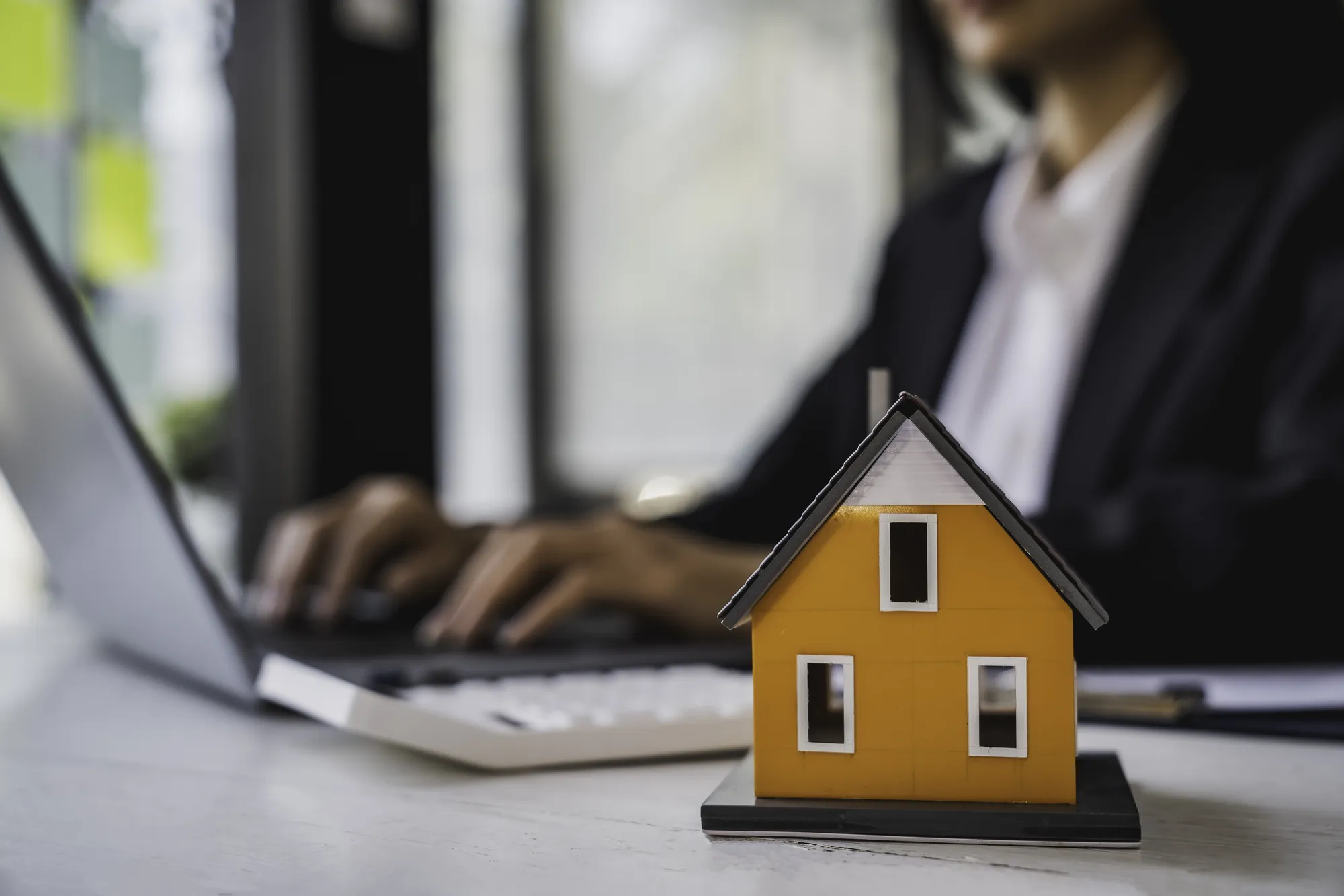 A woman is typing on a laptop with a small model house in front of her.