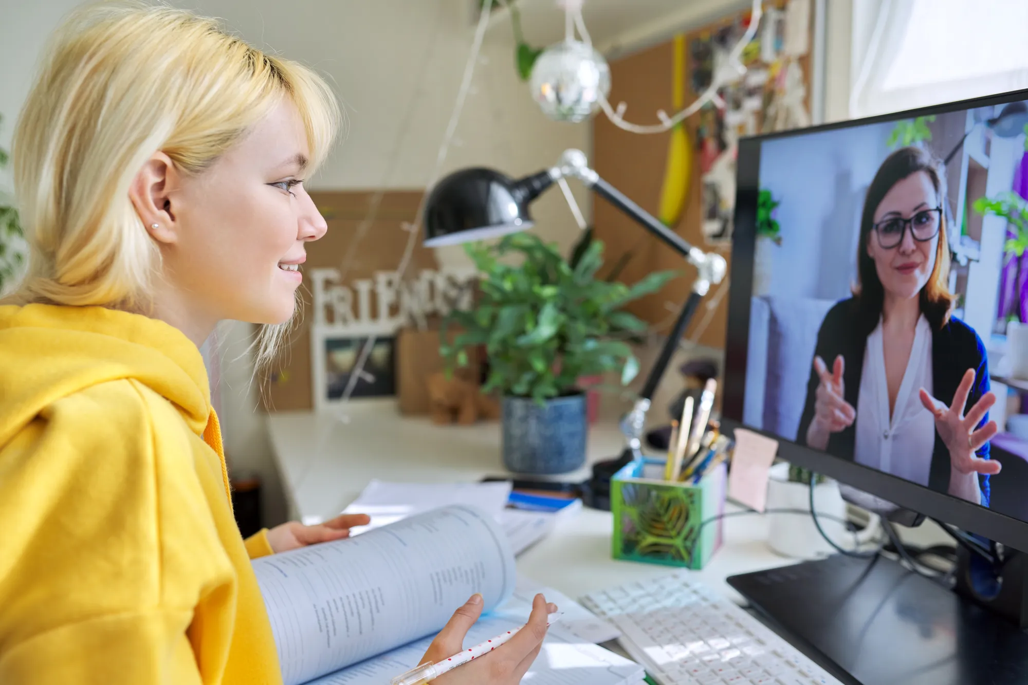 A woman with blonde hair and a yellow hoodie is sitting at a desk in front of a computer screen.