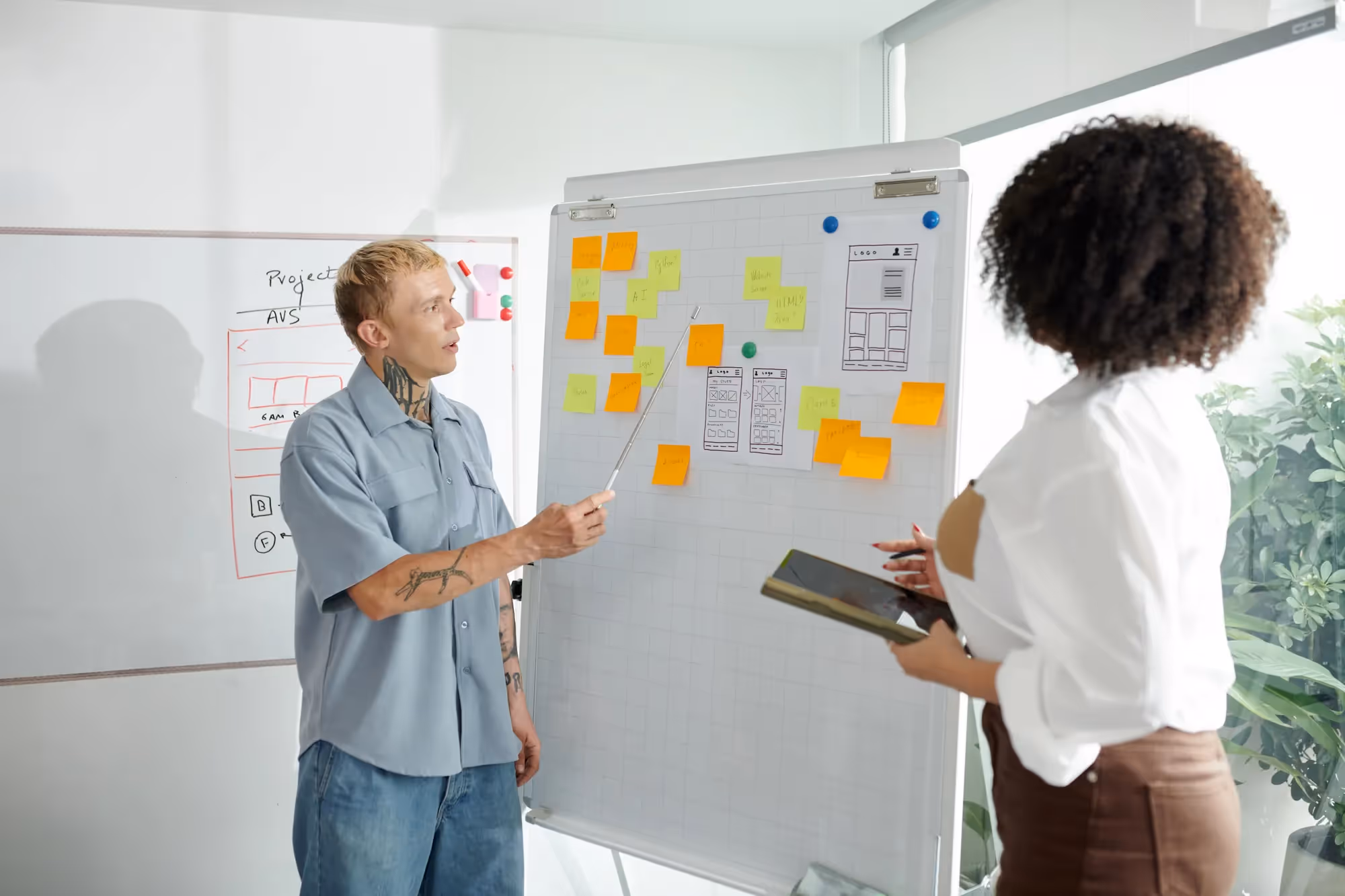 A man with a tattoo and a woman with a white shirt are standing in front of a white board with sticky notes on it.