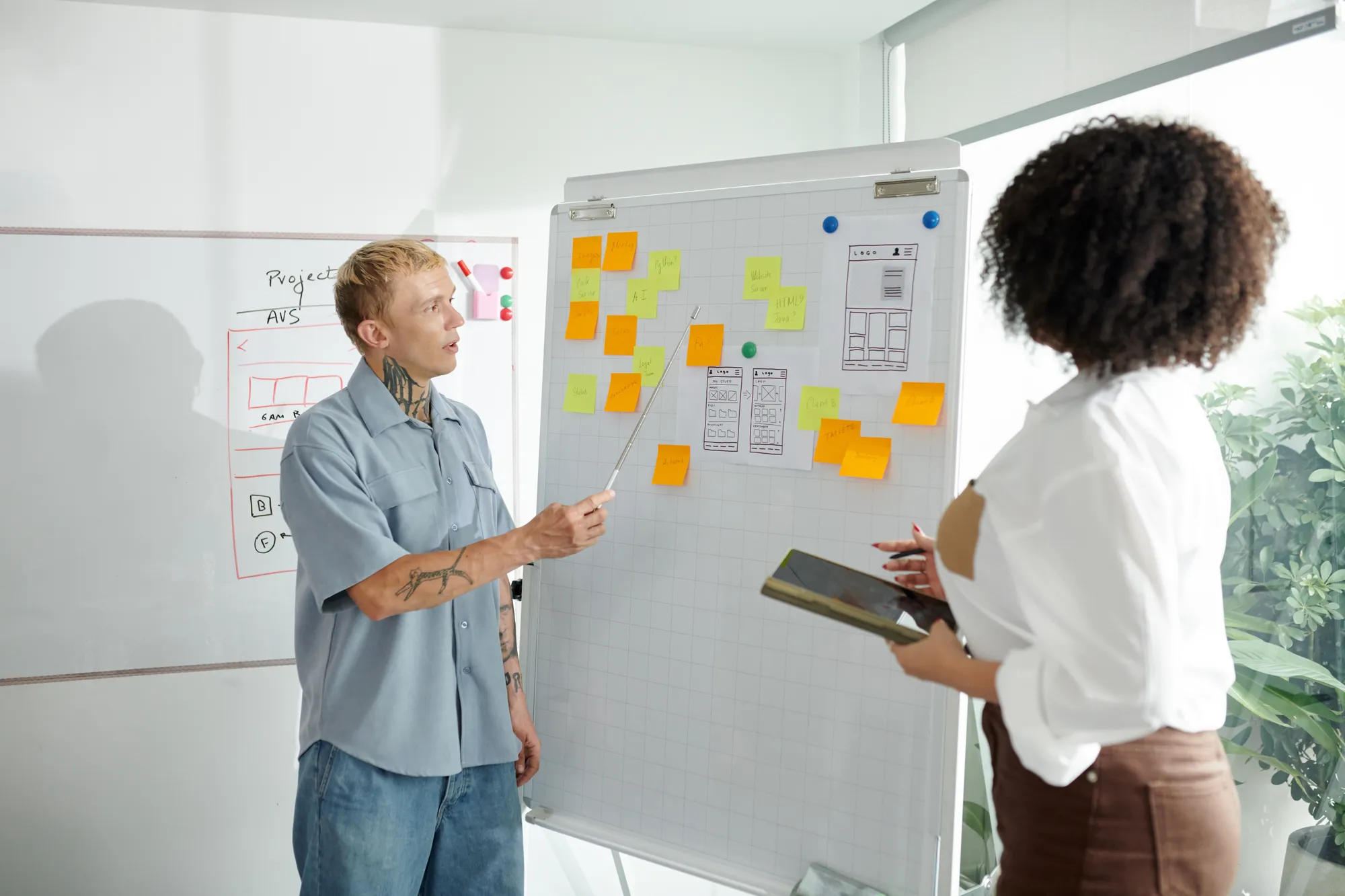A man with a tattoo and a woman with a white shirt are standing in front of a white board with sticky notes on it.