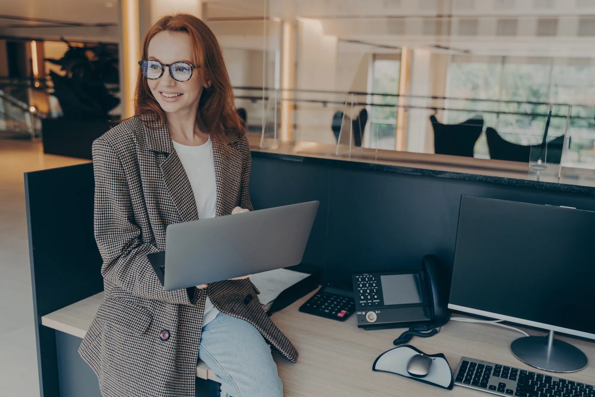 A woman in a brown coat is sitting at a desk with a laptop and a phone.