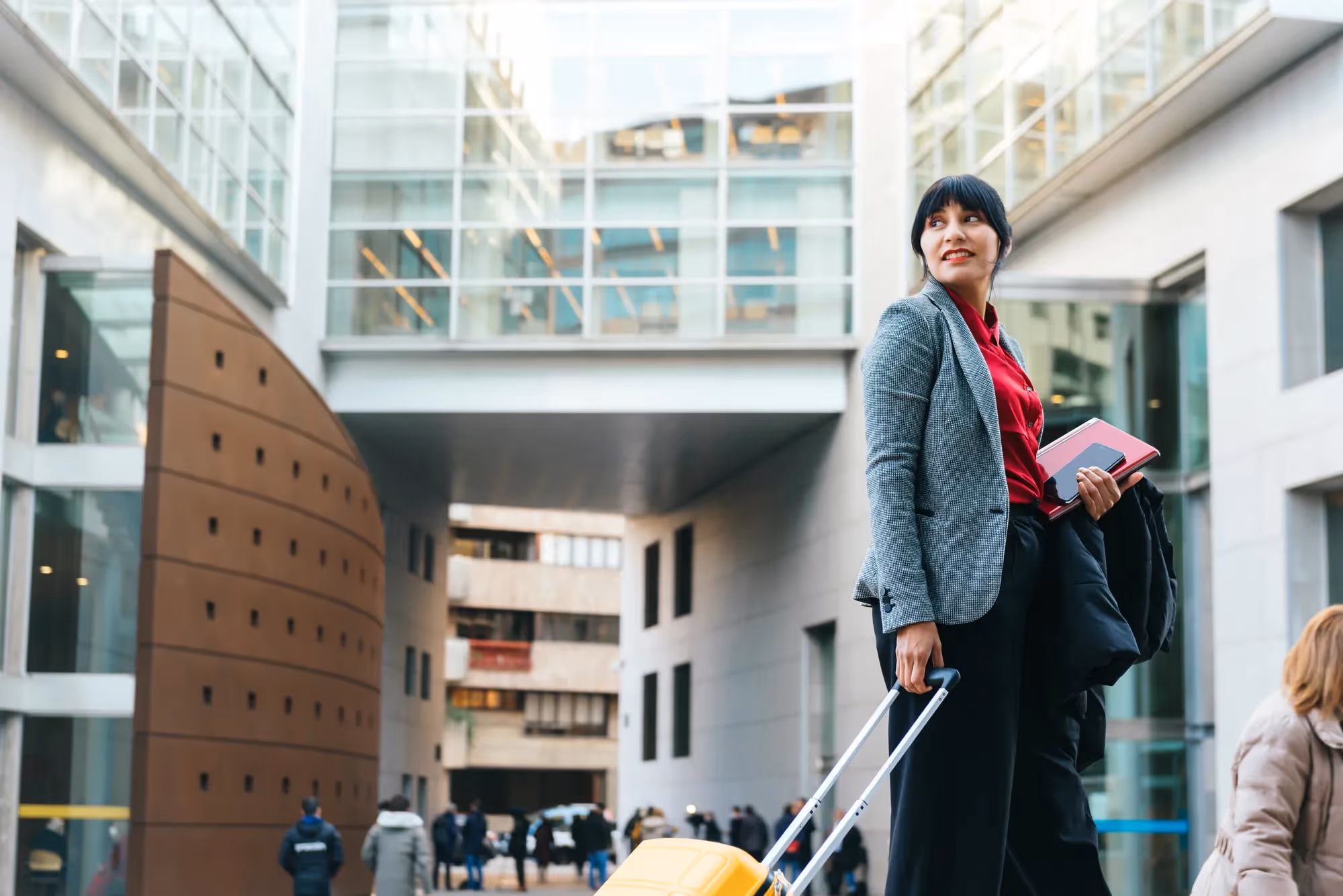 A woman in a grey suit is pulling a yellow suitcase.