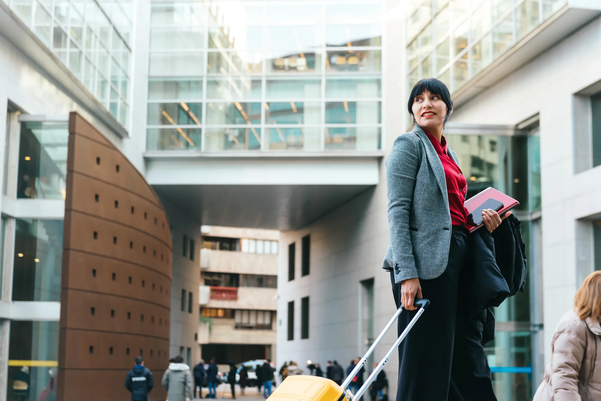 A woman in a grey suit is pulling a yellow suitcase.
