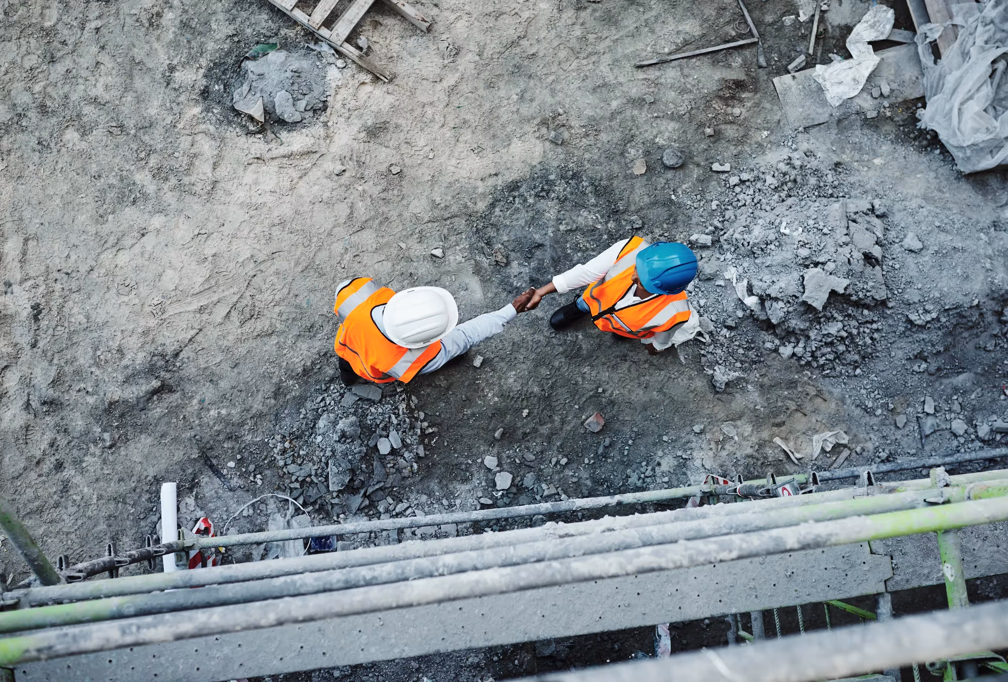 Two construction workers shake hands in front of a pile of rubble.