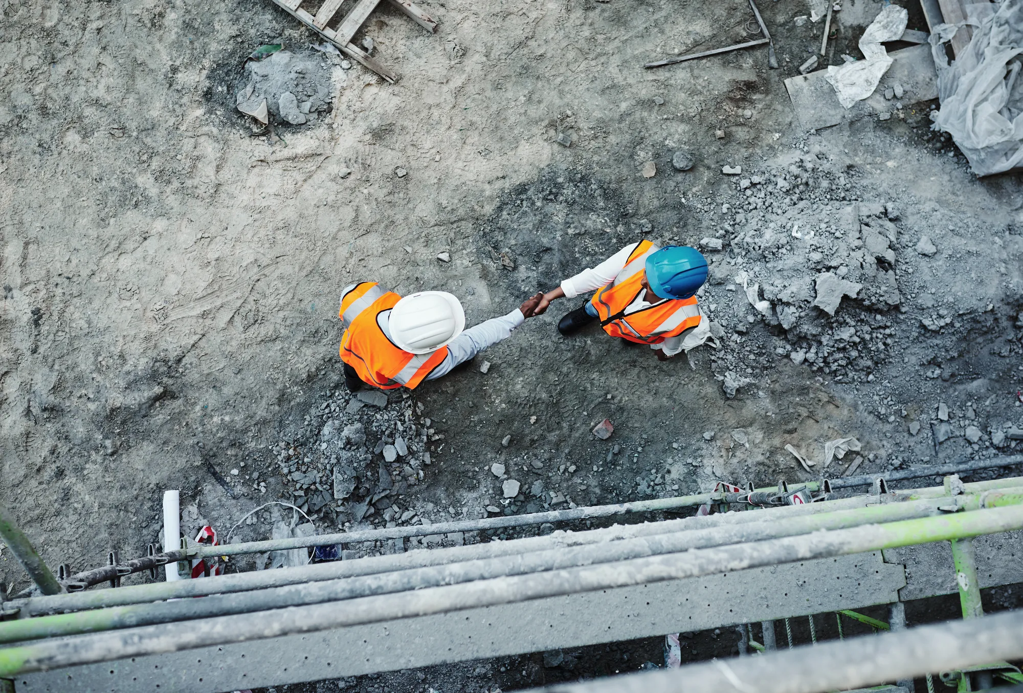 Two construction workers shake hands in front of a pile of rubble.