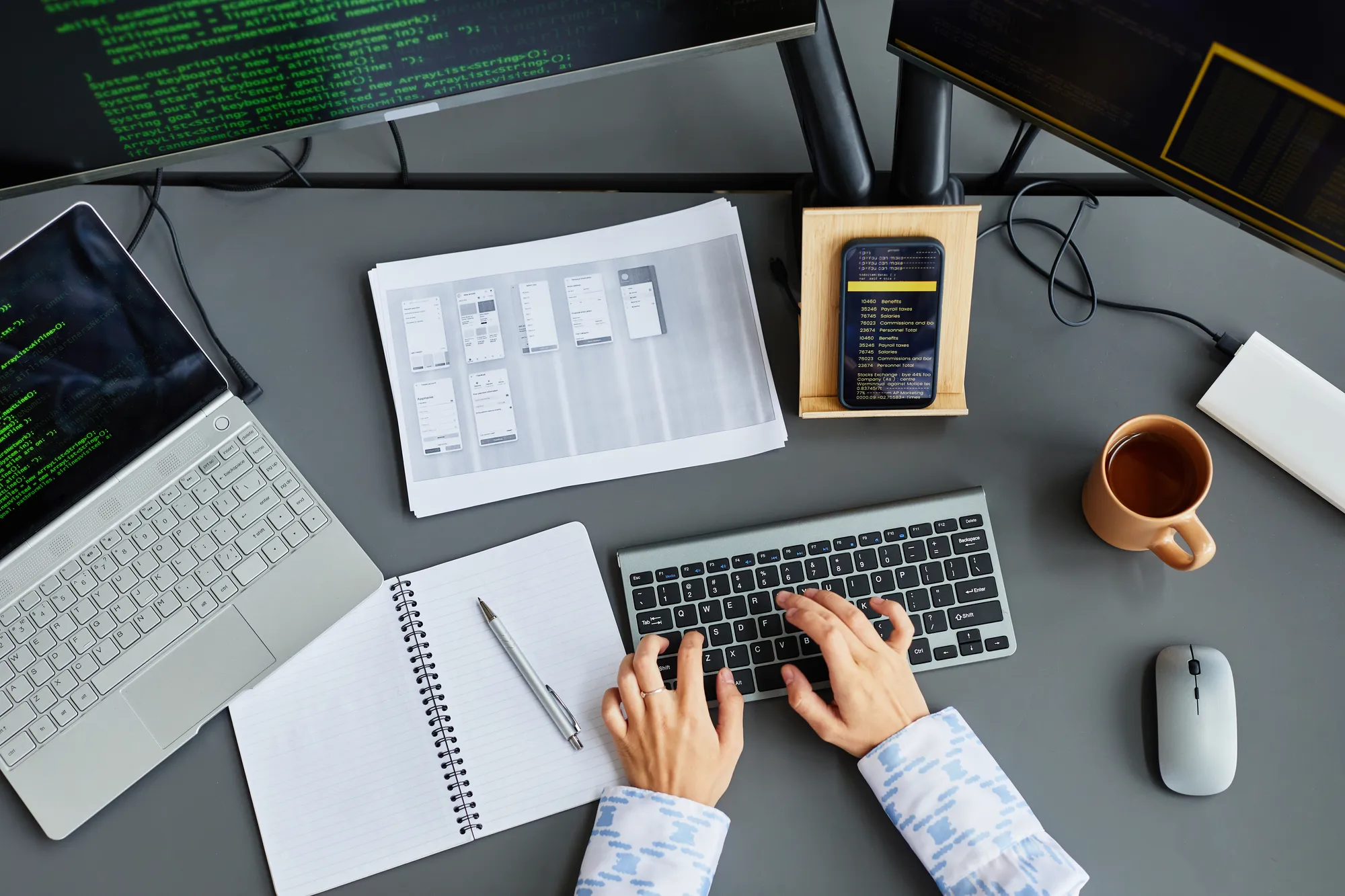 A person typing on a keyboard with a notebook and a cell phone on the desk.