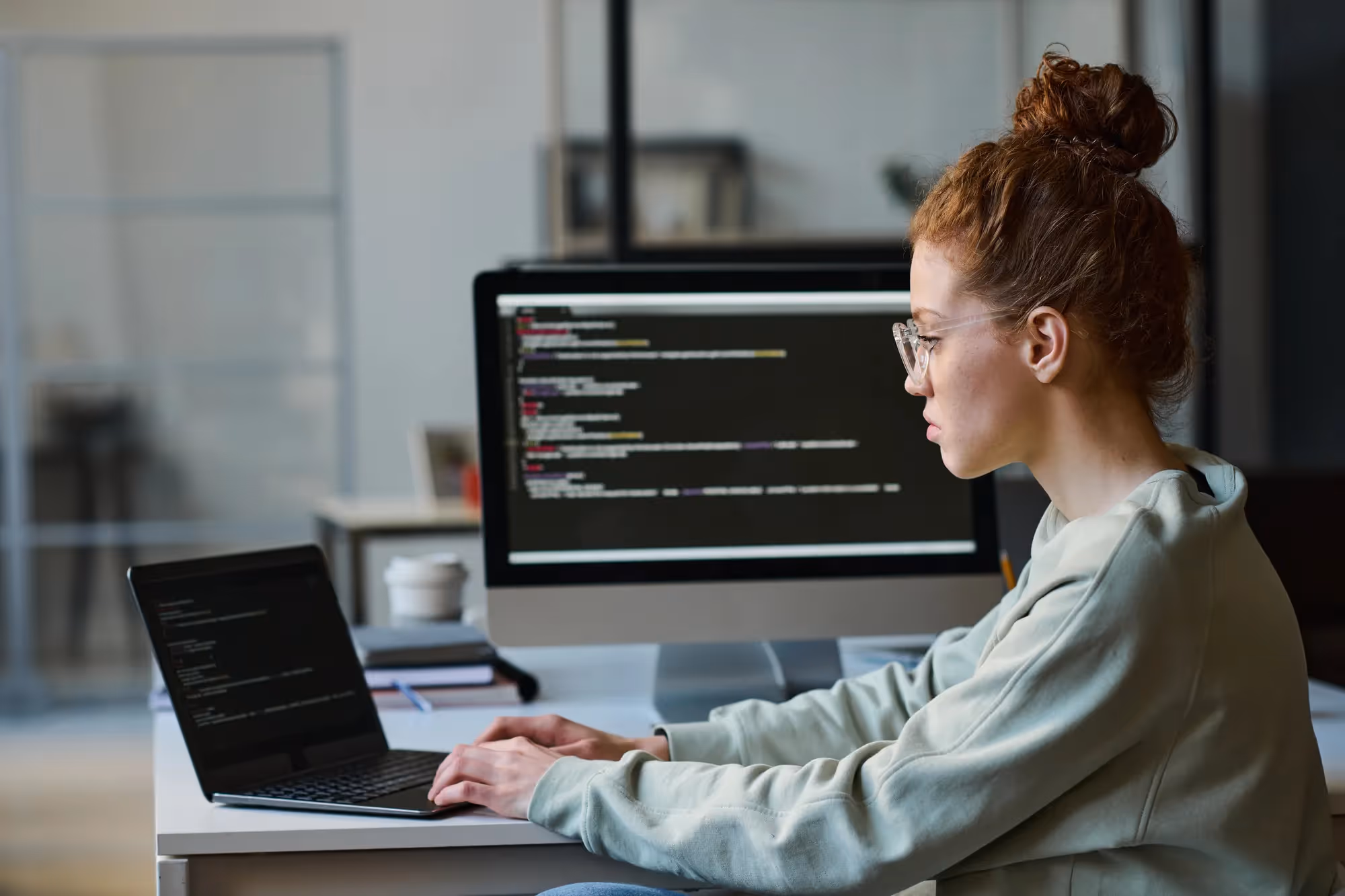 A woman is typing on a laptop in front of a computer monitor.