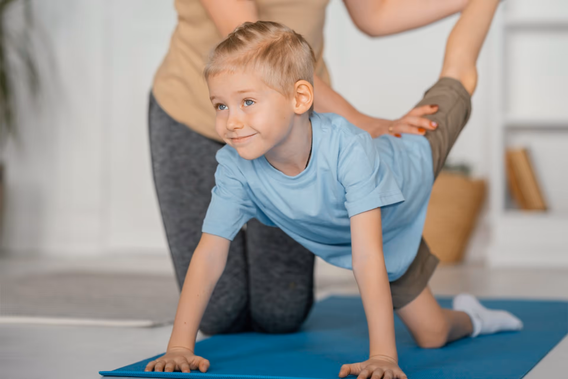 Ein Junge auf einer blauen Gymnastikmatte, der bei einer Dehnübung von einer Physiotherapeutin am Bein unterstützt wird.