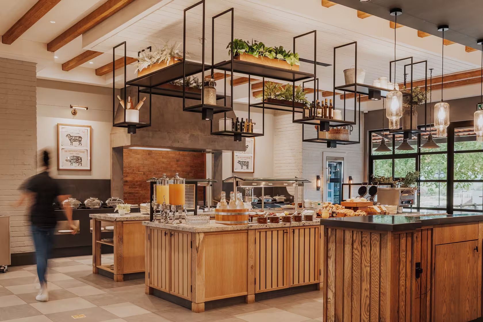 Modern buffet area with wooden counters, juice dispensers, shelves with plants, and a blurred person walking.