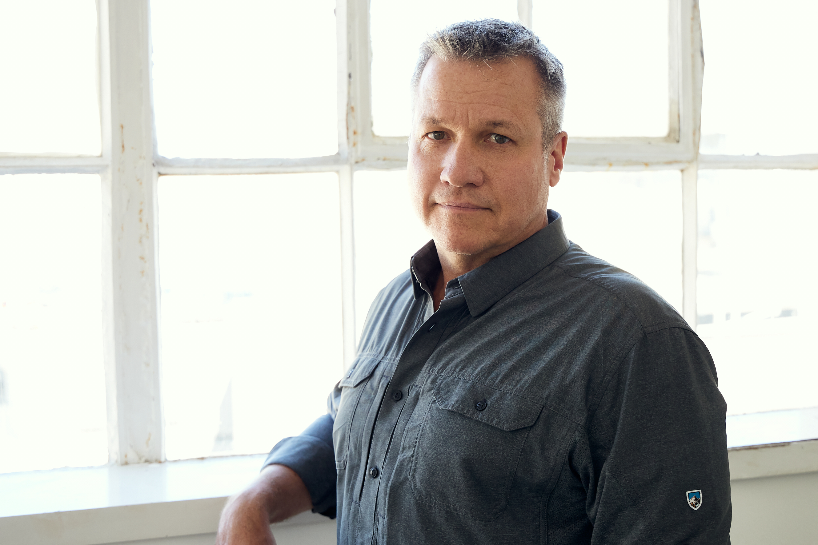 Man with gray hair wearing a dark buttoned shirt with sleeves rolled up, standing against gray backdrop.