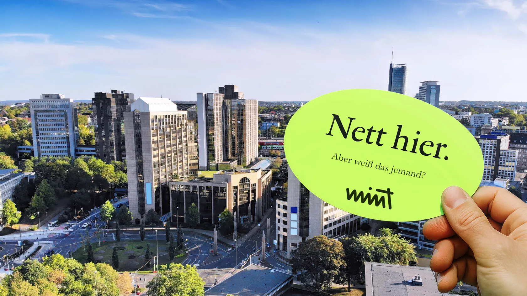 Picture of the city center of Essen, in front of it a hand holding a sticker that says “Nice here. But does anyone know that?” MINT