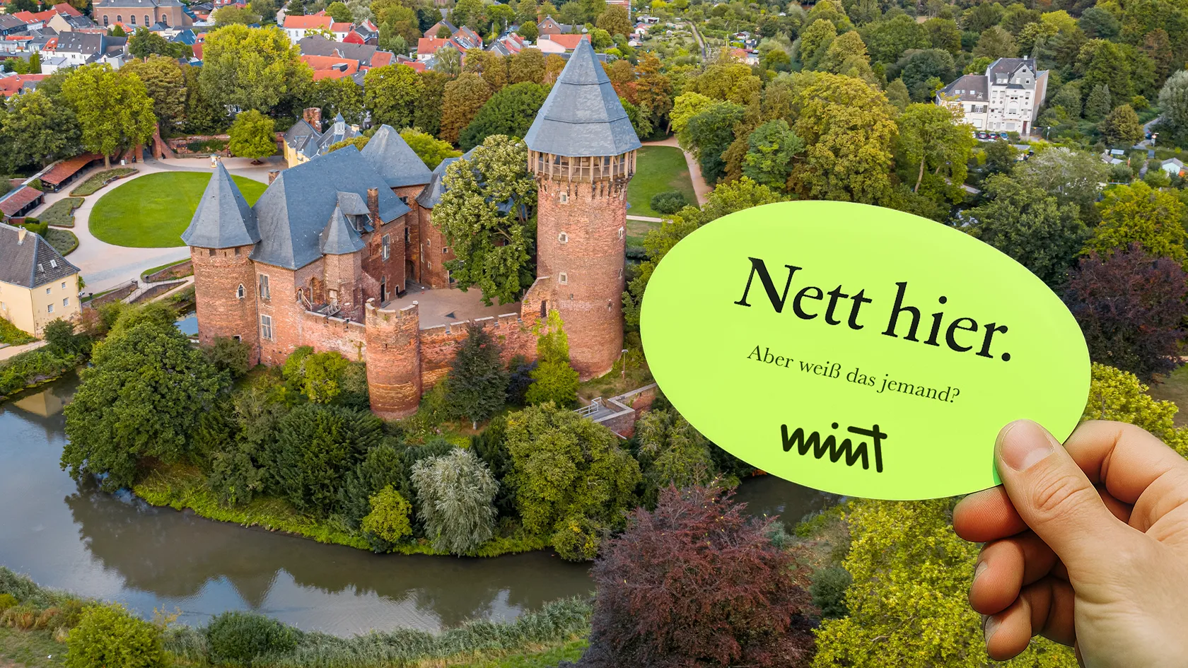 Bild der Burg Linn in Krefeld, davor eine Hand die einen Sticker hält auf dem steht "Nett hier. Aber weiß das jemand?" MINT