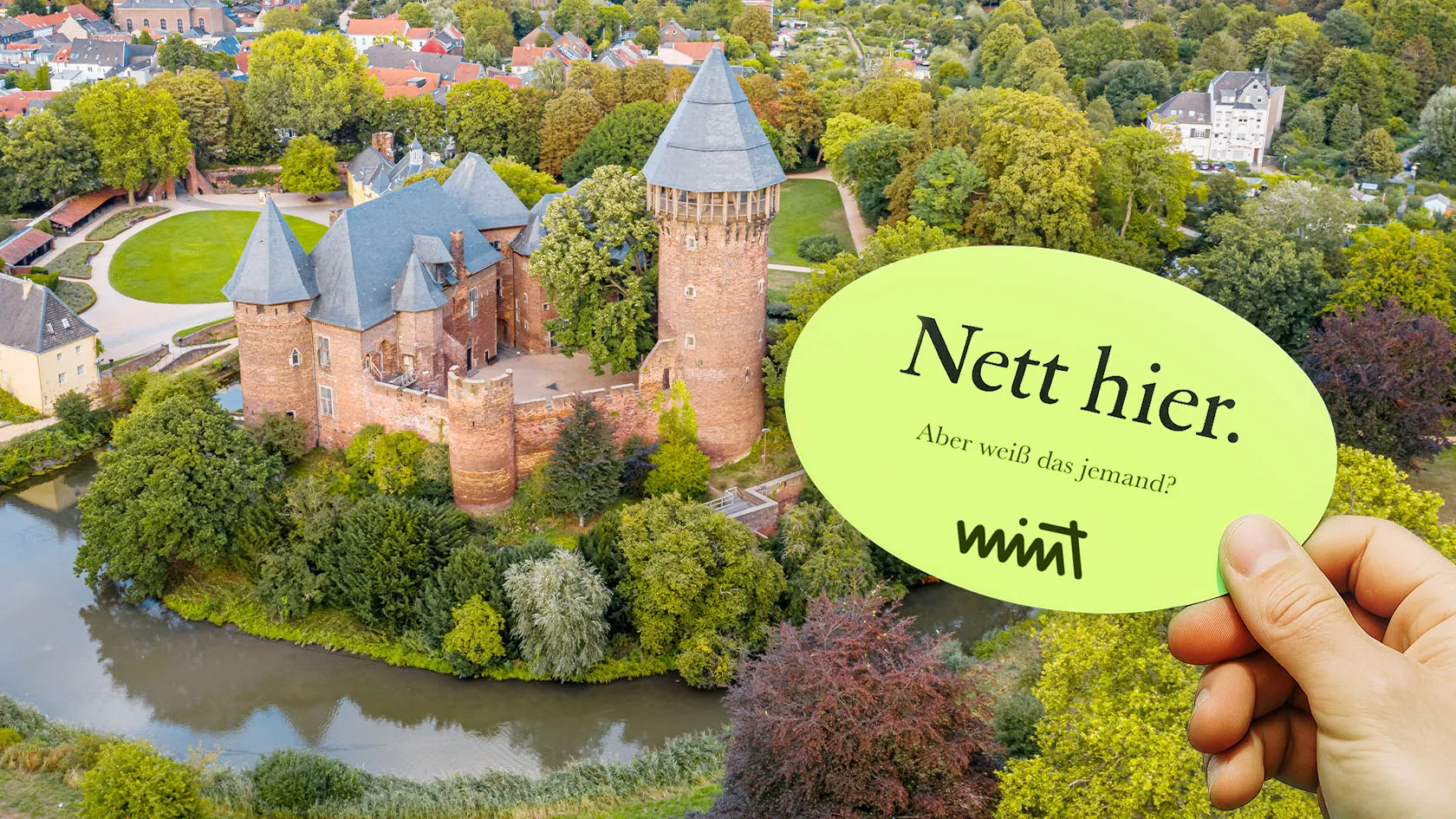 Bild der Burg Linn in Krefeld, davor eine Hand die einen Sticker hält auf dem steht "Nett hier. Aber weiß das jemand?" MINT
