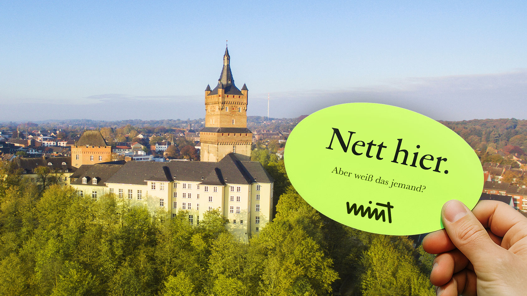 Bild der Schwanenburg in Kleve, davor eine Hand die einen Sticker hält auf dem steht "Nett hier. Aber weiß das jemand?" MINT