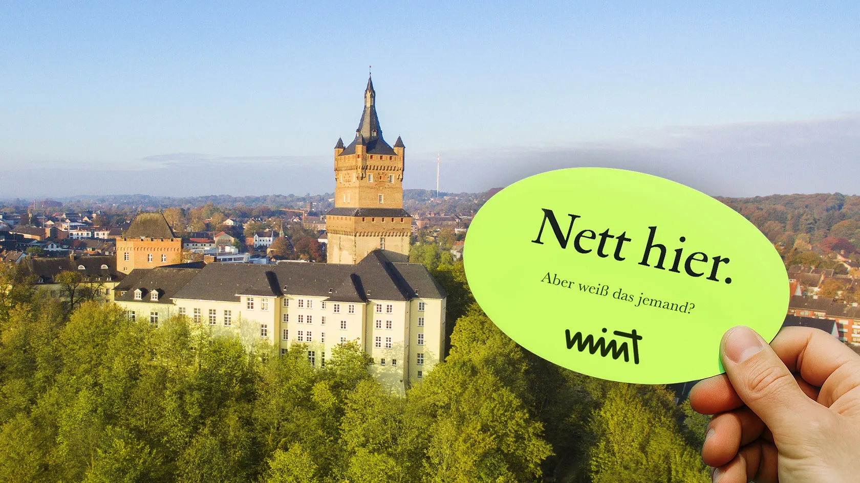 Bild der Schwanenburg in Kleve, davor eine Hand die einen Sticker hält auf dem steht "Nett hier. Aber weiß das jemand?" MINT