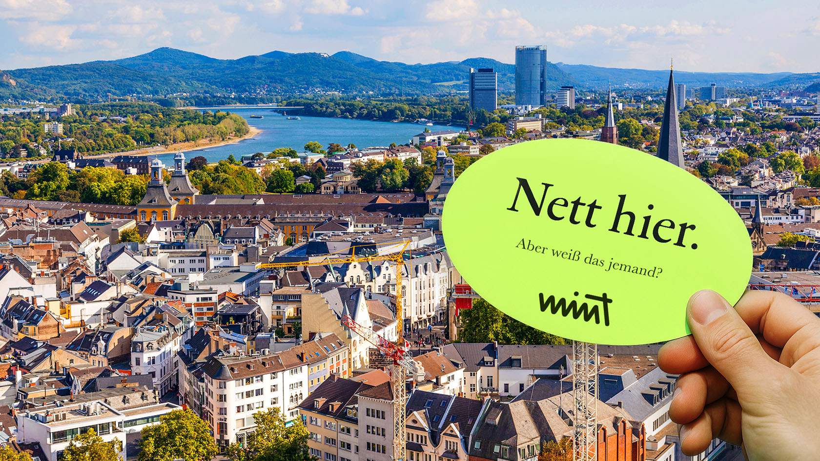 Bild des Stadtzentrums von Bonn mit Blick auf den Rhein, davor eine Hand die einen Sticker hält auf dem steht "Nett hier. Aber weiß das jemand?" MINT