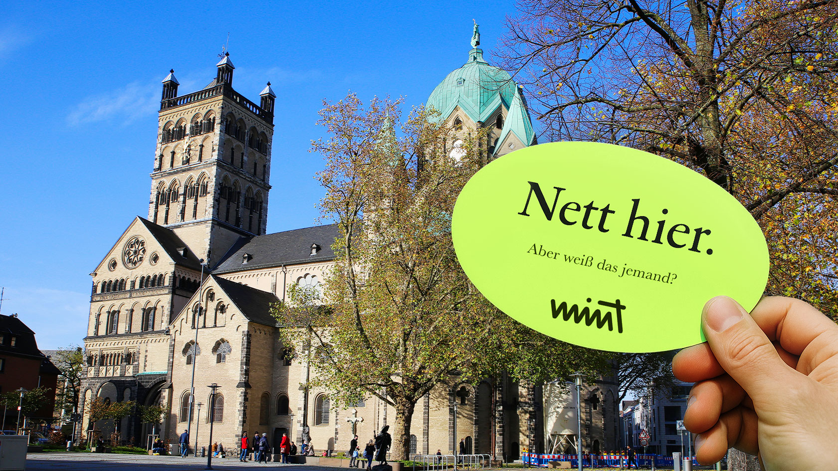 Bild des Stadtzentrums von Neuss, davor eine Hand die einen Sticker hält auf dem steht "Nett hier. Aber weiß das jemand?" MINT