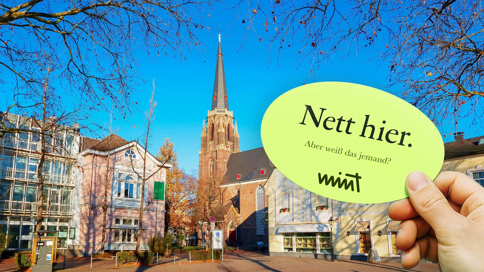 Picture of the city center of Moers, in front of it a hand holding a sticker that says “Nice here. But does anyone know that?” MINT