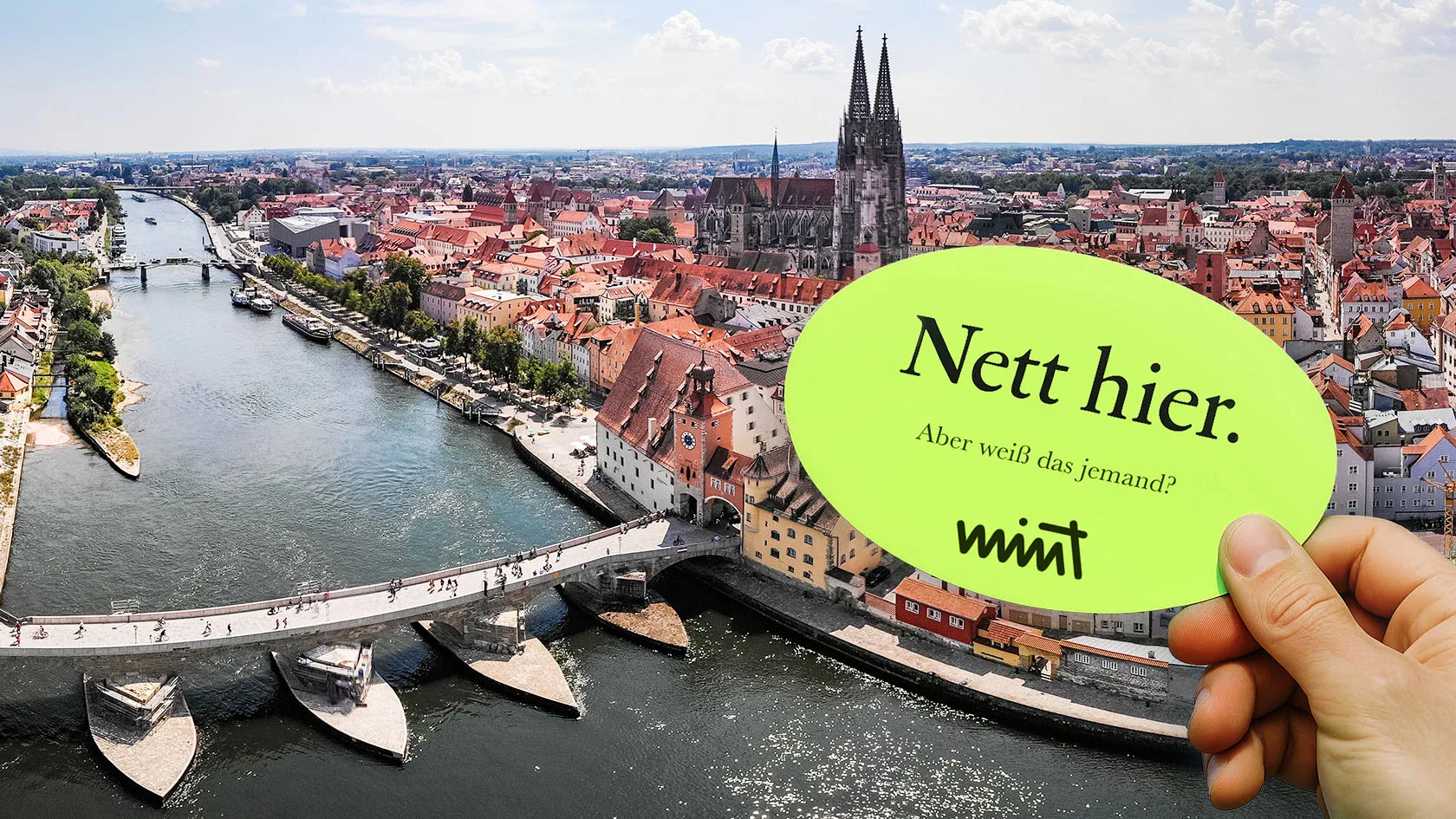 Bild der Stadt Regensburg am Flussarm, davor eine Hand die einen Sticker hält auf dem steht "Nett hier. Aber weiß das jemand?" MINT