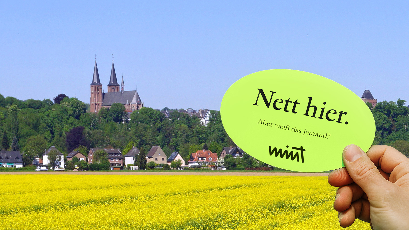 Bild mit einem Rapsfeld im Vordergrund, im Hintergrund sieht man ein Dorf, davor eine Hand die einen Sticker hält auf dem steht "Nett hier. Aber weiß das jemand?" MINT