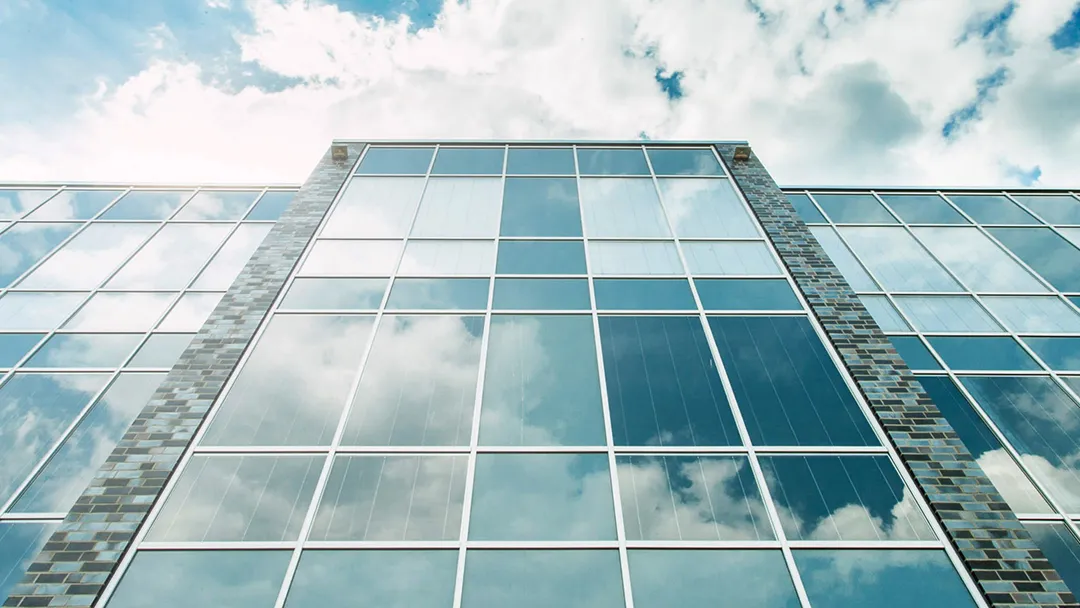 Facade view of a modern building with large glass windows reflecting the sky with clouds.