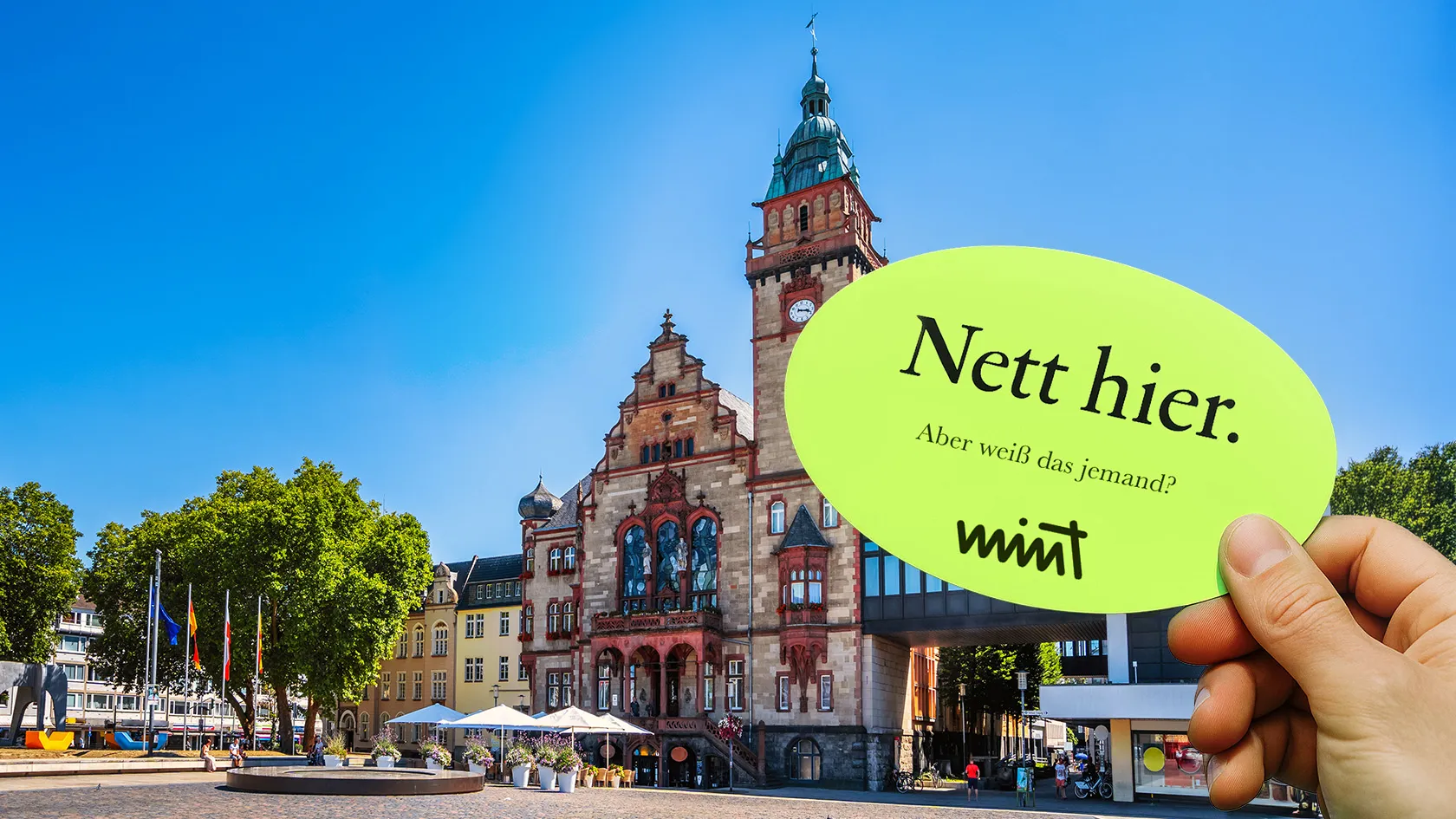 Bild von einem Marktplatz in Mönchengladbach, , davor eine Hand die einen Sticker hält auf dem steht "Nett hier. Aber weiß das jemand?" MINT