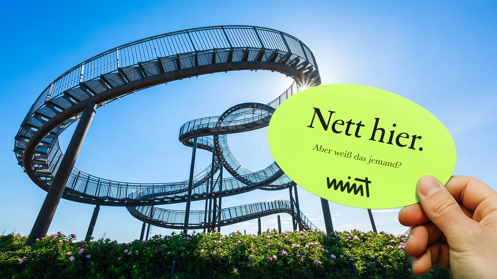 Picture of the tourist attraction “Tiger and Turtle” in Duisburg, in front of it a hand holding a sticker that says “Nice here. But does anyone know that?” MINT