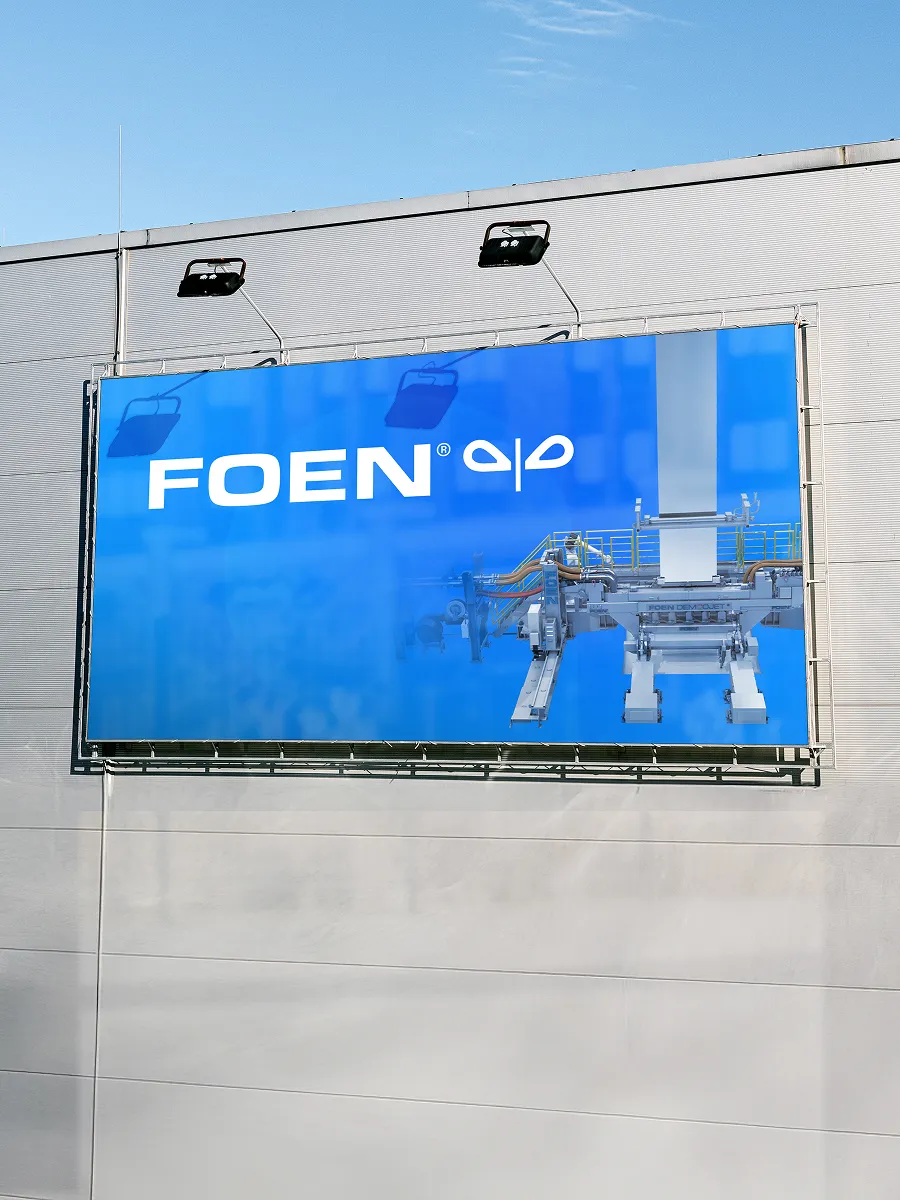Außenwand eines Gebäudes mit einer großen blauen Werbetafel, die das FOEN-Logo und eine industrielle Maschine zeigt.