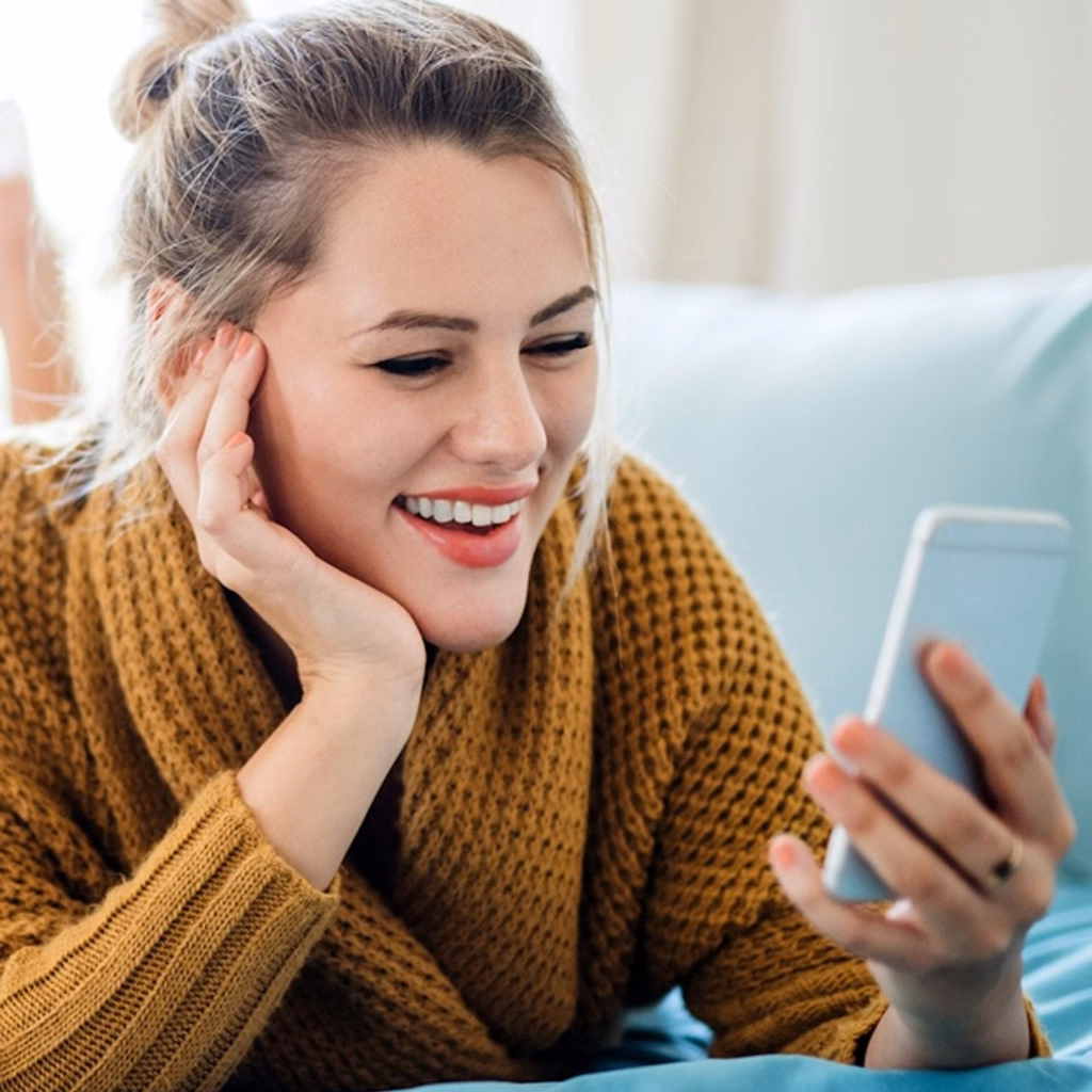 Smiling woman in yellow sweater lying on couch looking at smartphone in her hand.