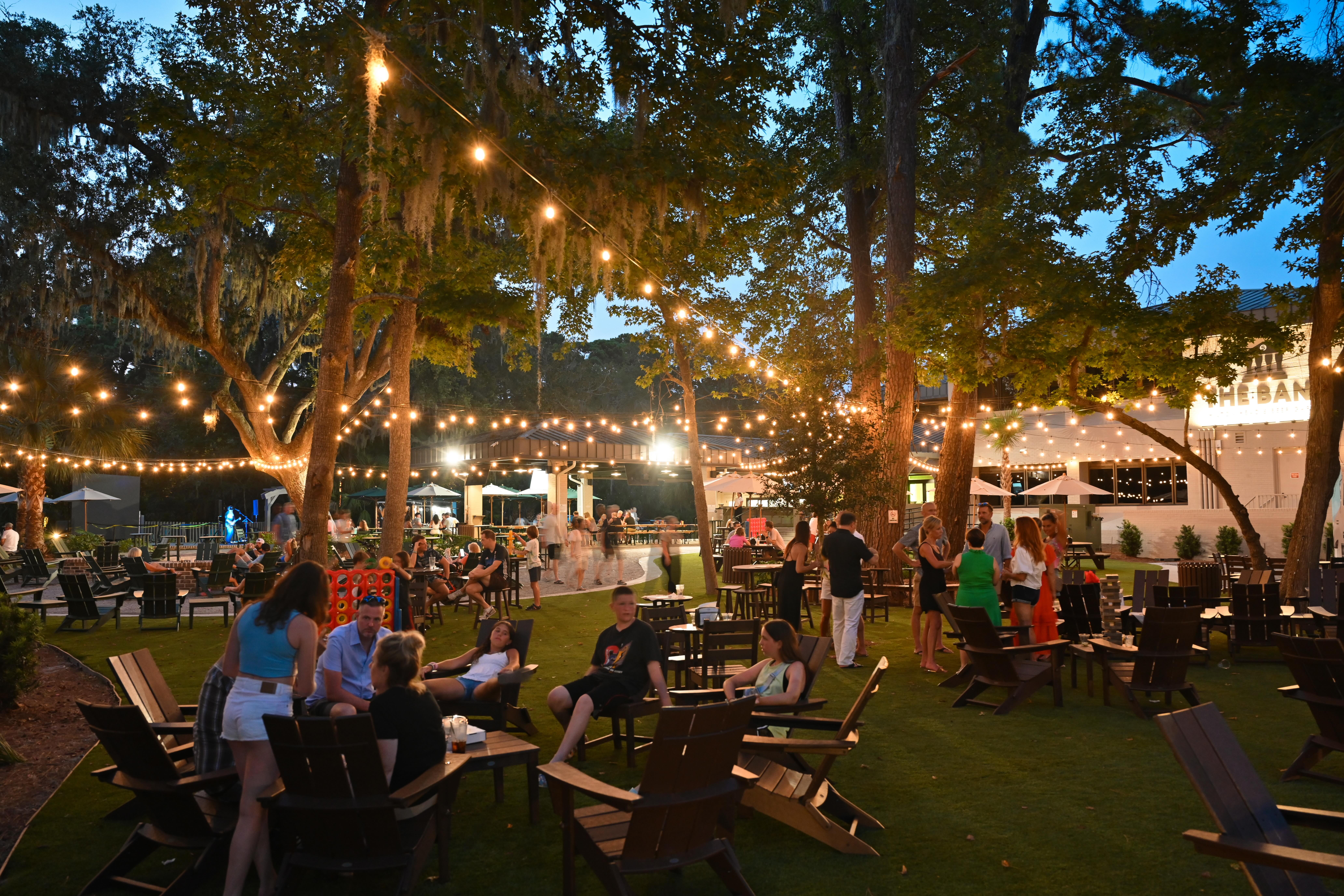 Outdoor evening scene of people socializing under string lights in a wooded seating area with Adirondack chairs and tables.