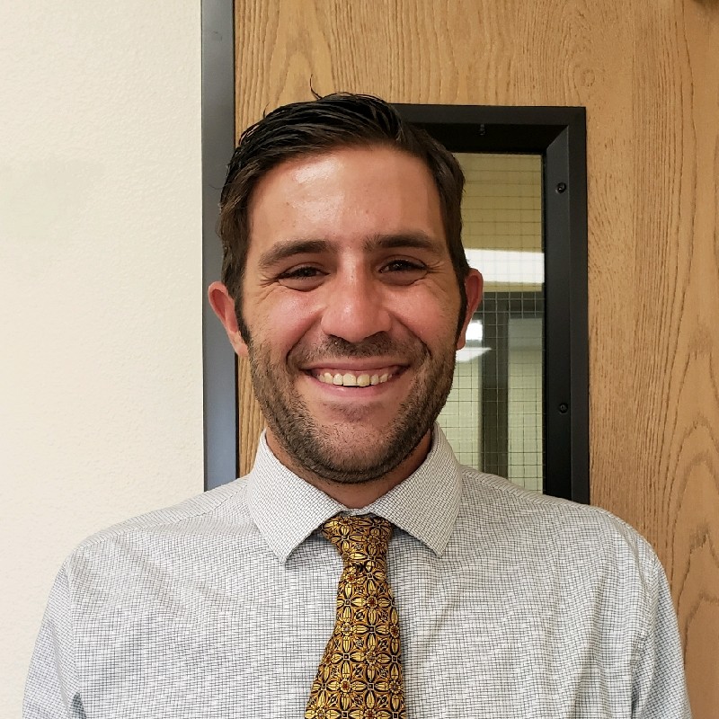 Smiling man with short dark hair wearing a patterned light gray shirt and yellow patterned tie in front of a wooden door.