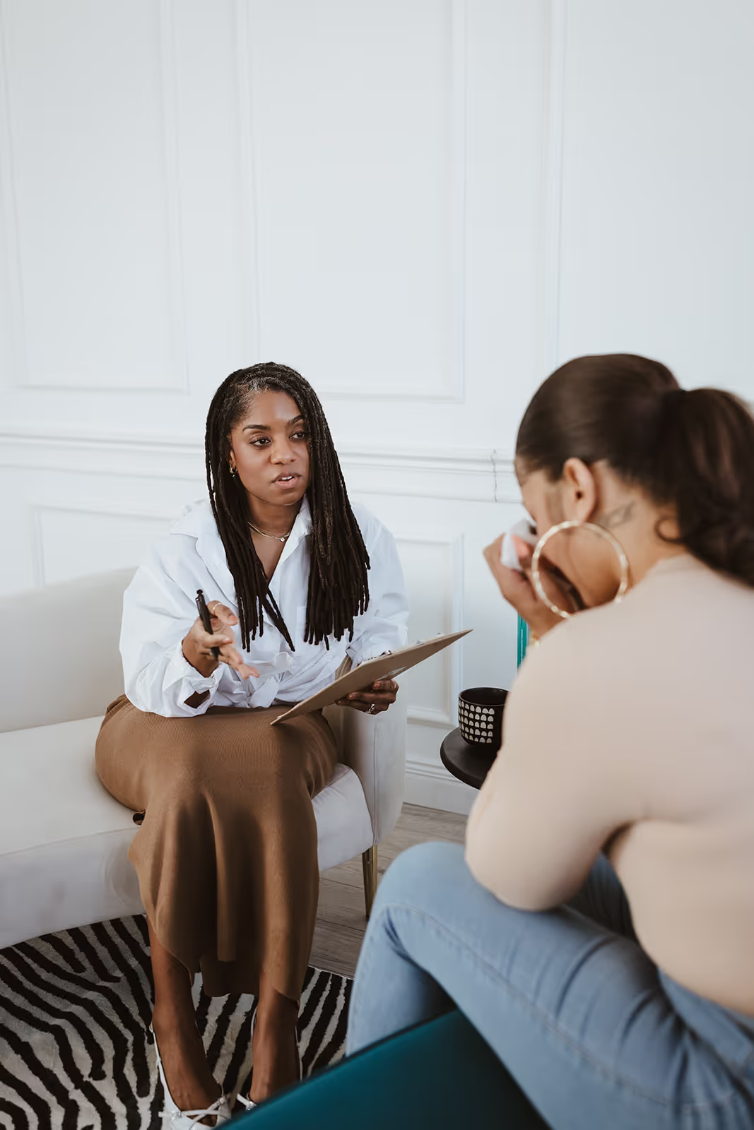 Brittany L. Maxshure, a Black woman with long locs, sits on a cream-colored couch, attentively engaging with a client during a therapy session. She wears a white blouse and brown skirt, holding a clipboard and pen, while the client, a woman in a beige top and blue jeans, wipes tears with a tissue. The setting is warm and modern, featuring a zebra-patterned rug, a black coffee table with a patterned mug, and a light, neutral-toned background.