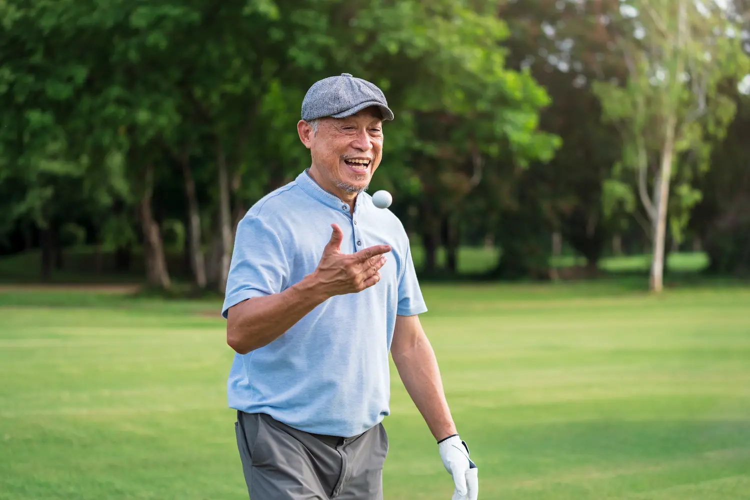 Active older man smiling and looking in the distance on a golf green tossing a golf ball up in the air from his hand.