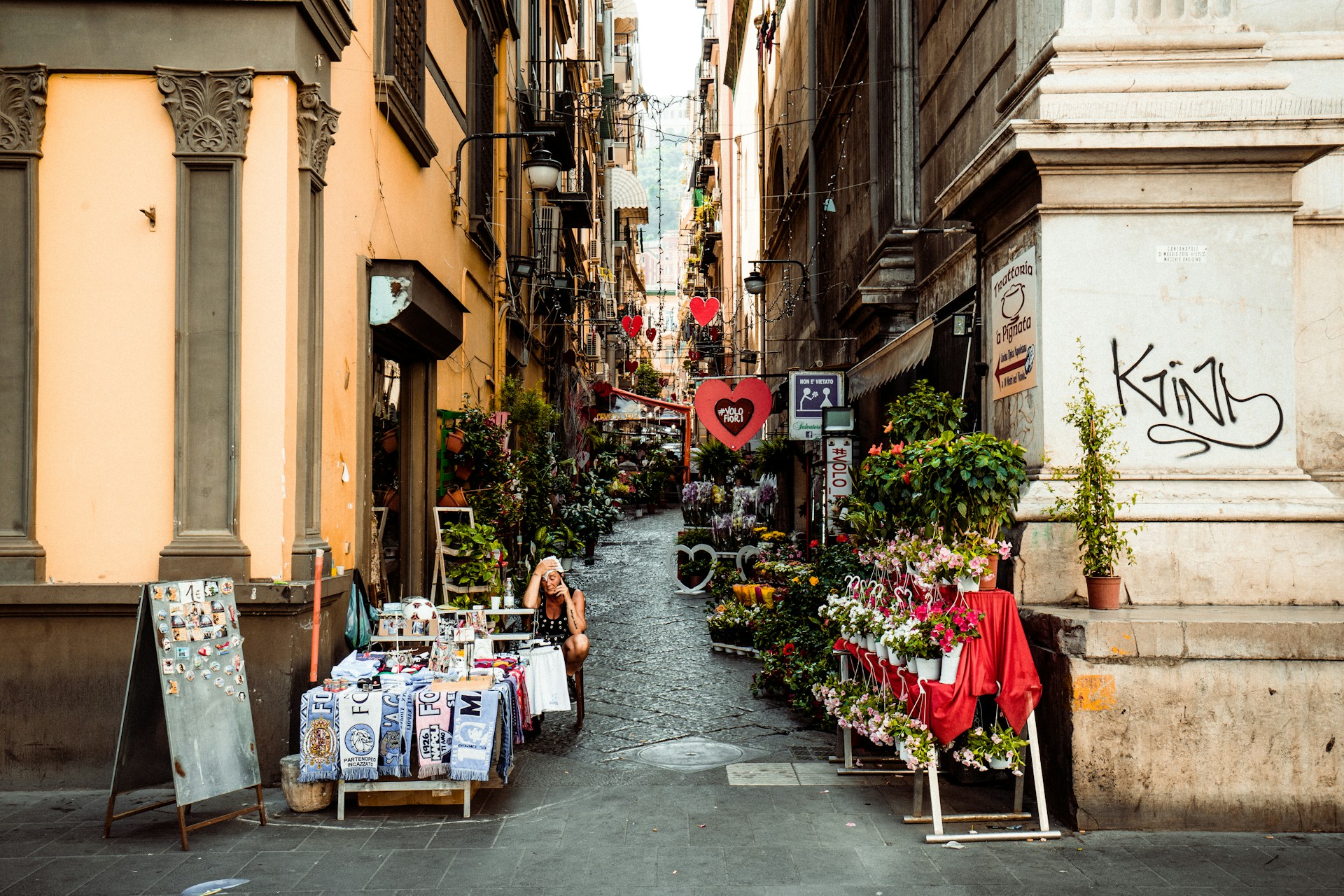 A narrow alleyway in Naples, Italy, adorned with vibrant flowers and lush plants lining the cobblestone path.