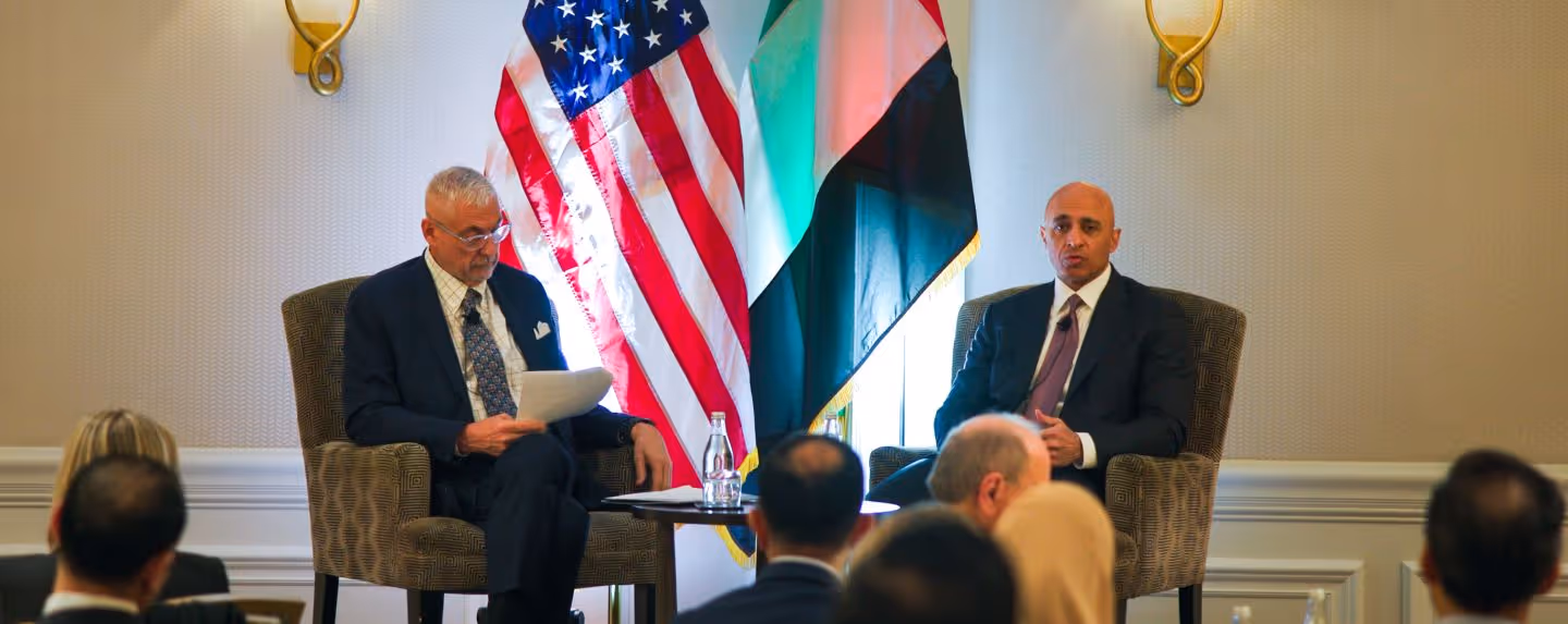 Two speakers sit on stage in armchairs during a formal event, with the U.S. and UAE flags displayed behind them. One man reads from notes while the other speaks, as an audience listens in the foreground.
