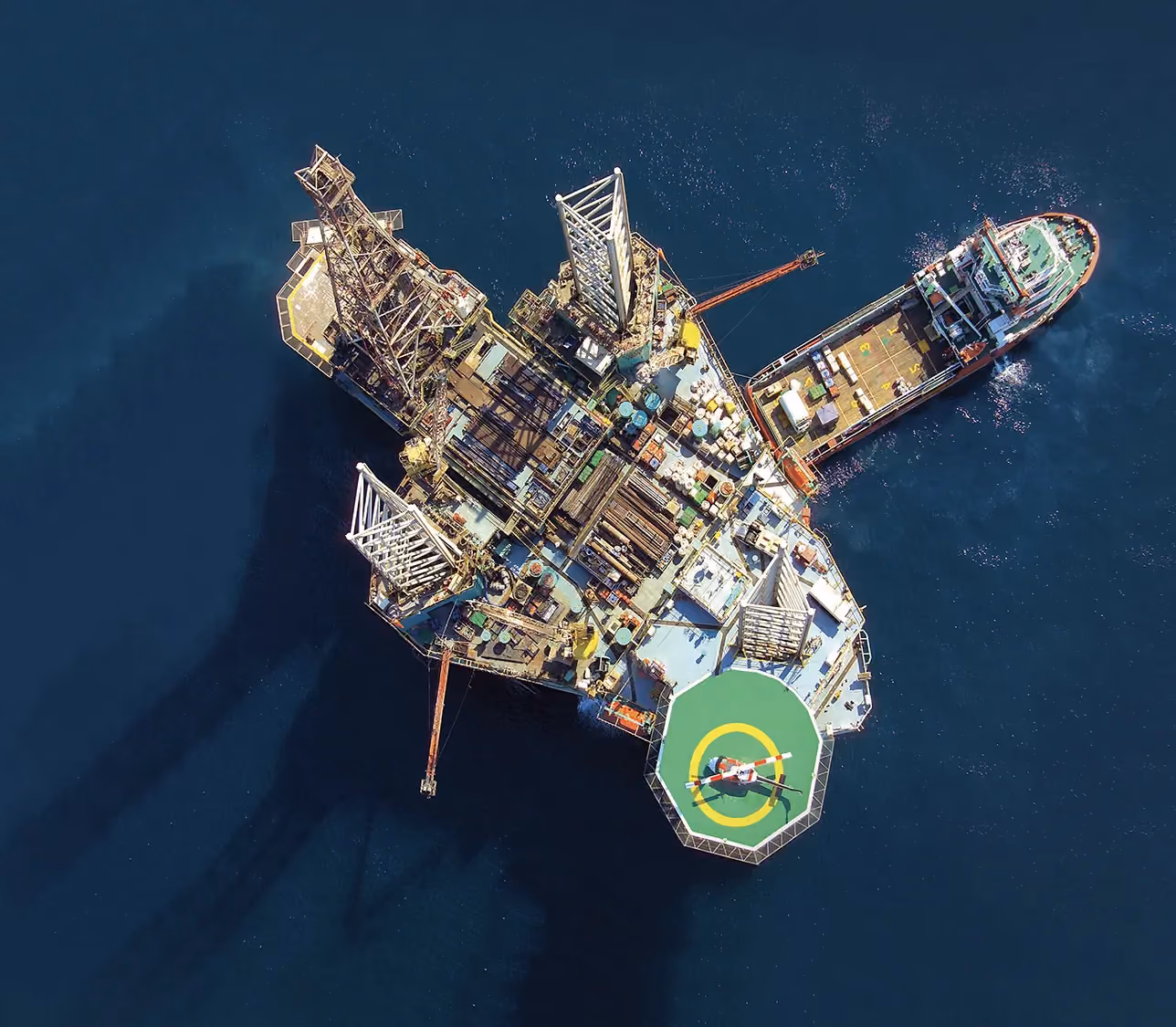 Aerial view of an offshore drilling rig with a supply vessel alongside and a helicopter on the helipad.
