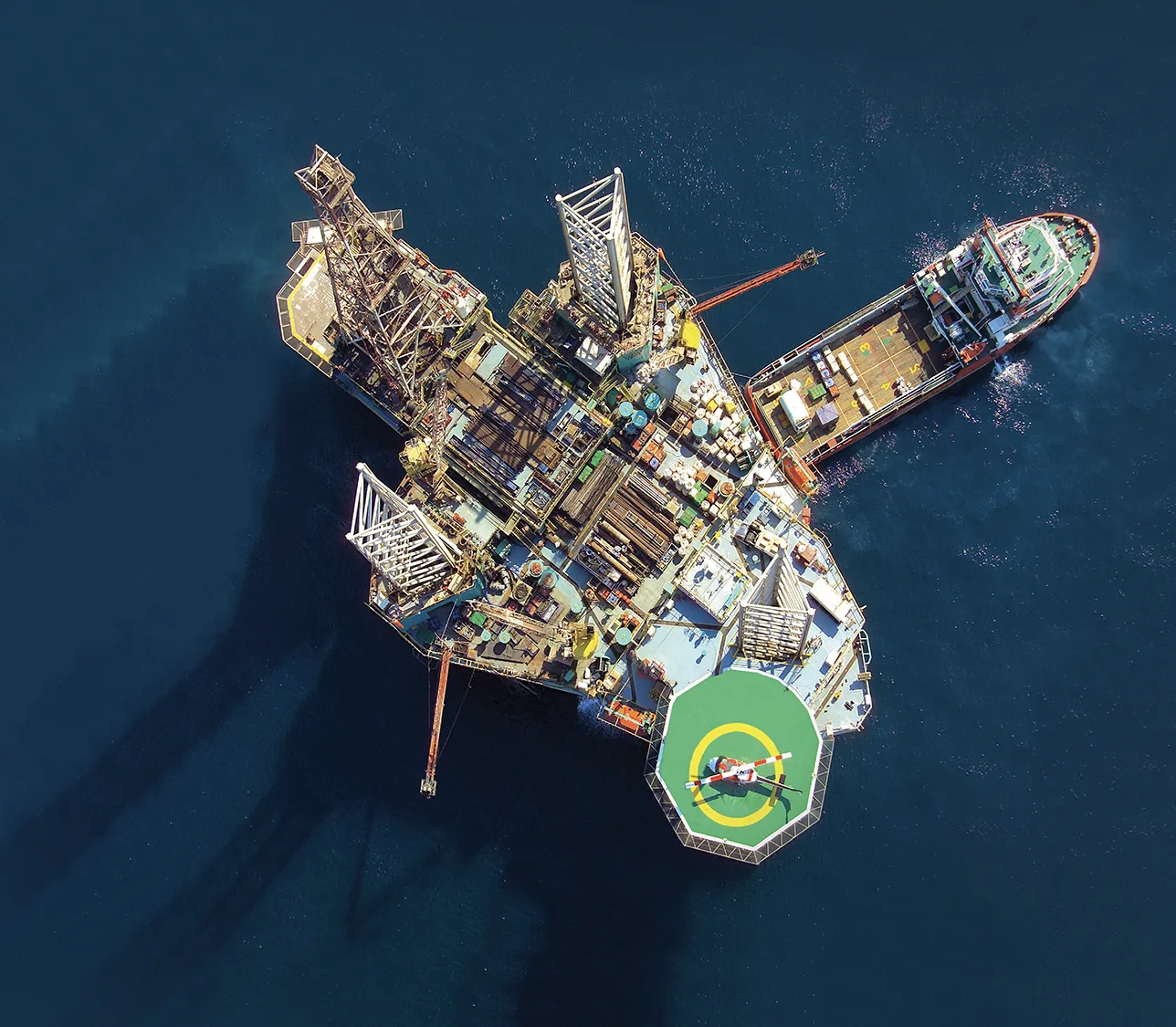 Aerial view of an offshore drilling rig with a supply vessel alongside and a helicopter on the helipad.