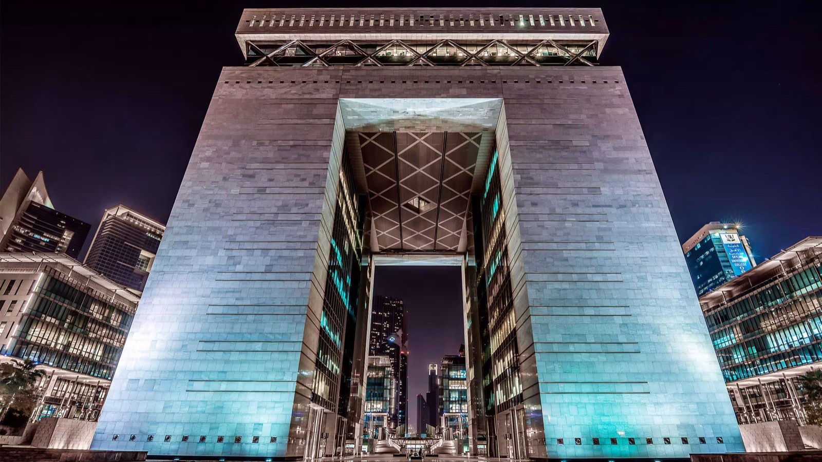 Night view of the DIFC Gate building in Dubai, lit in teal against a dark sky.