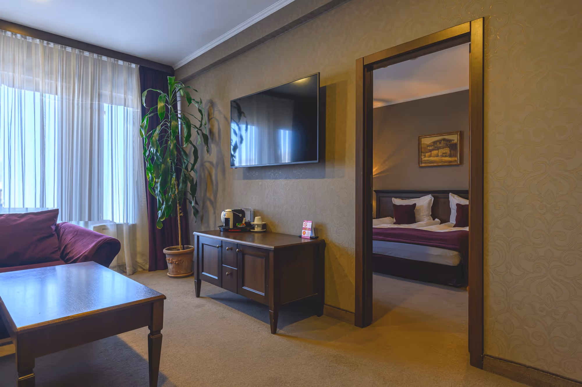 Просторен и елегантен хол в апартамент ВИП, обзаведен с удобни дивани в бордо, стилна дървена маса, мек мокет и тежки златисти завеси, създаващи уютна и луксозна атмосфера.