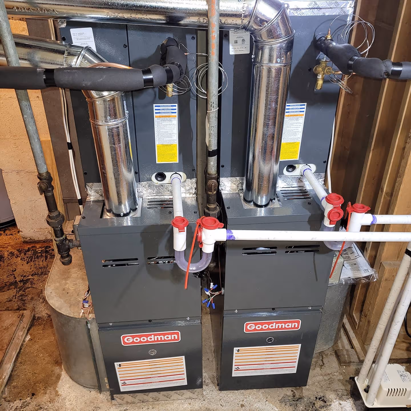 Two Goodman furnaces with metal pipes in a basement utility room