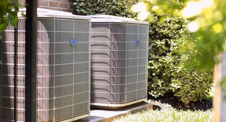 Two air conditioning units sitting outside near green bushes and brick wall