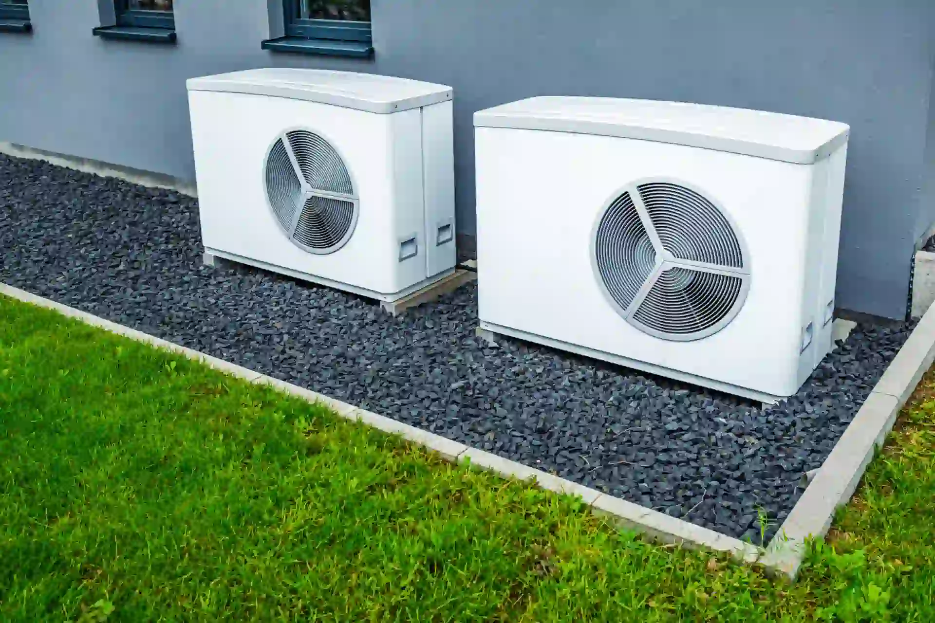 Two white air source heat pump units on gray rocks near green grass