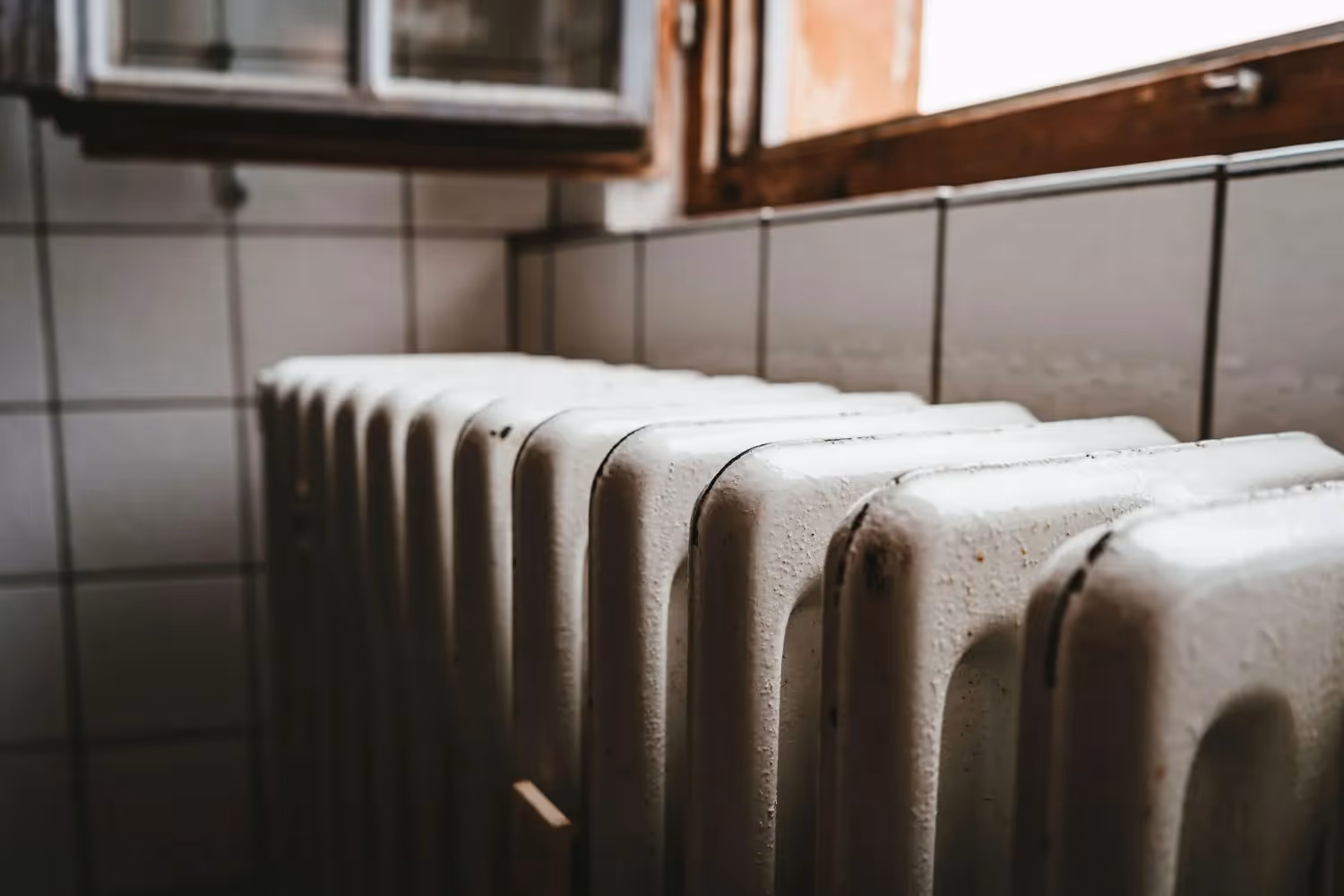 Old white radiator with vertical fins against tiled wall near window