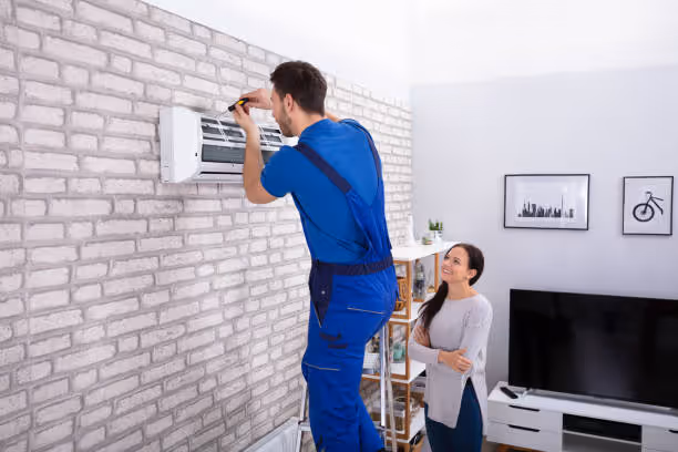 Technician in blue uniform installing air conditioner on white brick wall