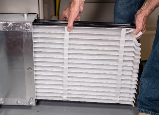 Hands holding a white pleated air filter next to metallic unit