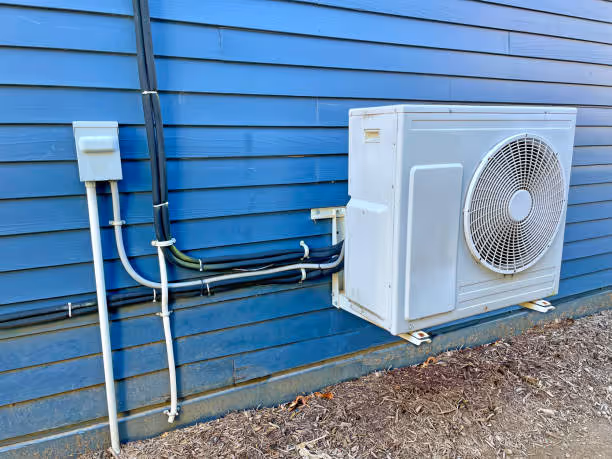 White air conditioning unit mounted on blue wooden siding with electrical conduits