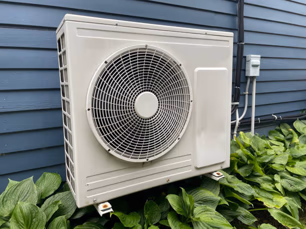 White air conditioning unit mounted on blue siding near green plants