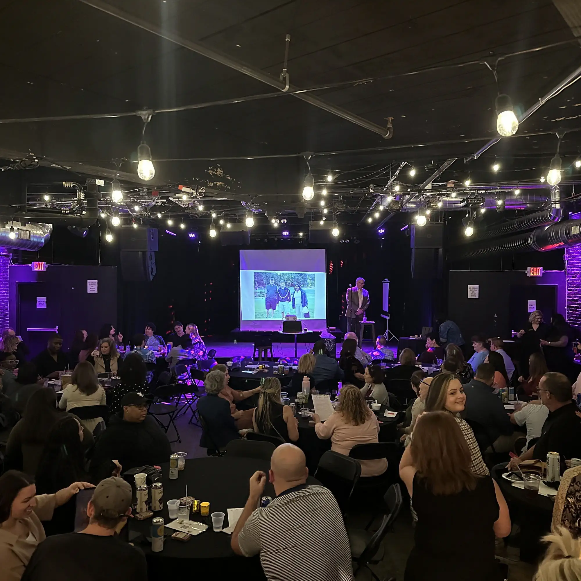 Guests seated at tables facing a presentation on the stage screen at Taffeta Music Hall